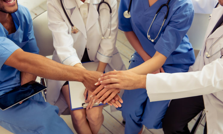 Cropped image of medical doctors of different nationalities and genders holding hands together and smiling, sitting in office