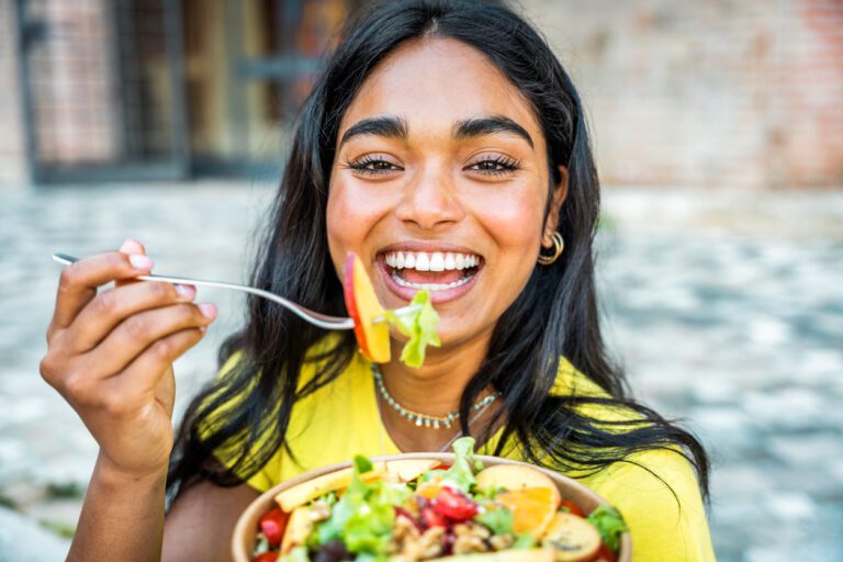 Portrait of young happy african woman eating healthy salad sitting on city street with green fresh ingredients Asian black student girl having vegan vegetarian lunch break outdoors